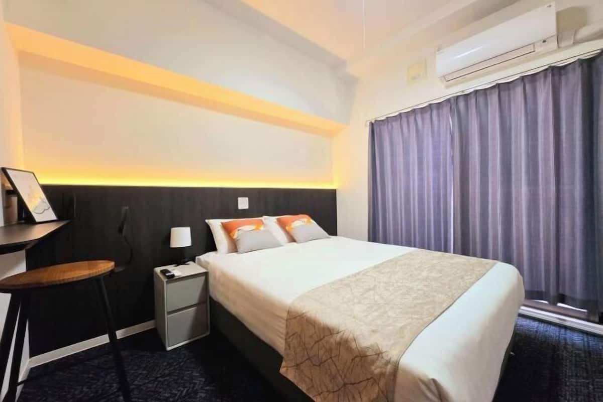 A hotel room with a bed, bedside table, lamp, and an air conditioning unit.
