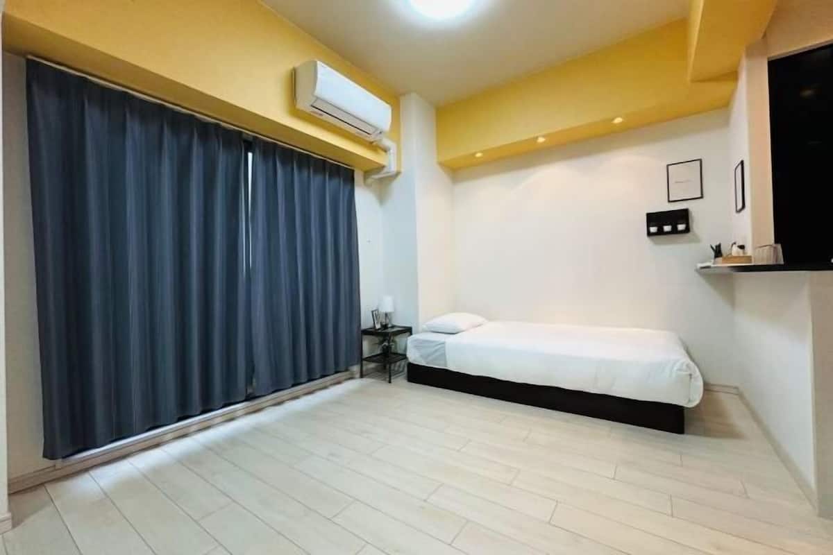 A small, modern bedroom with a single bed, a nightstand, a wall-mounted air conditioner, and a large window with curtains.