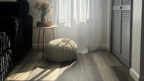 Ein Zimmer mit einer schwarzen Couch, einem runden Hocker, einem kleinen Tisch mit einer Vase und einem Fenster mit durchsichtigen Vorhängen.