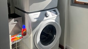Eine gestapelte Waschmaschine und ein Trockner, zusammen mit einer Flasche Tide Waschmittel und einer Packung Bounce Textilerfrischer auf einem Regal.