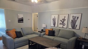 A modern living room with a grey sofa, a coffee table, and a patterned throw. There are framed cactus prints on the wall and a lamp on a side table. - Cowboy Casa - DFW, AT&T Stadium, Stockyards (Hurst)