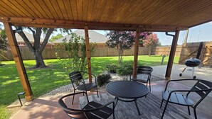 A covered patio with a round table and four chairs, a wooden pergola overhead, and a backyard with a lawn and a wooden fence. - Cowboy Casa - DFW, AT&T Stadium, Stockyards (Hurst)