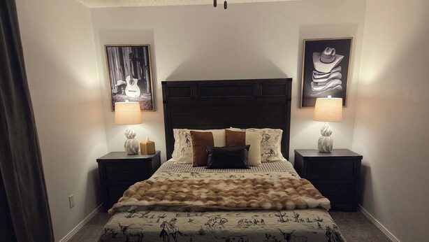 A bedroom with a large bed, two bedside tables with lamps, a headboard, and two wall-mounted pictures. - Cowboy Casa - DFW, AT&T Stadium, Stockyards (Hurst)
