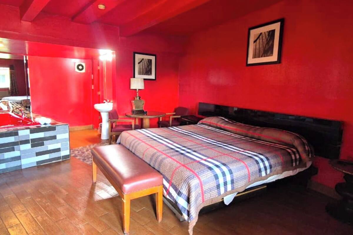 A red-themed bedroom with a large bed, a bench, a small table with a lamp, and a mirror.