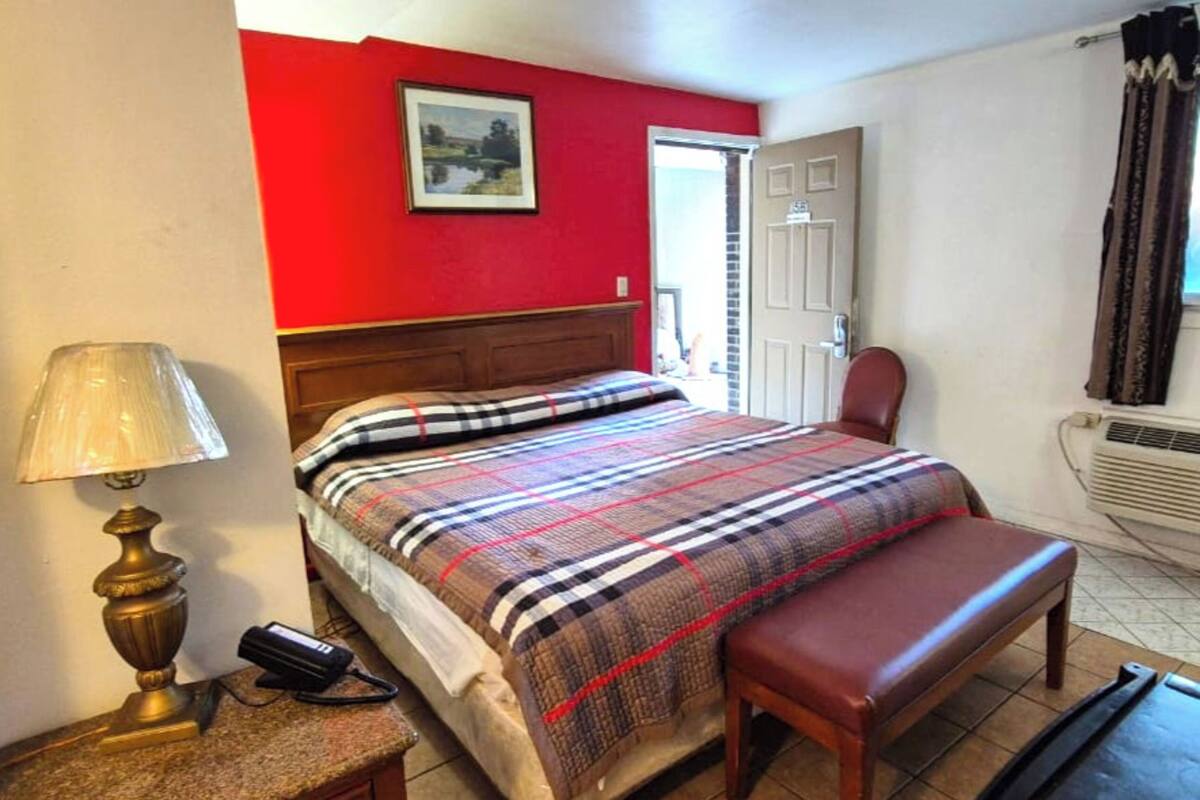 A bedroom with a bed, a bedside lamp, a bench, a picture on the wall, and a door.