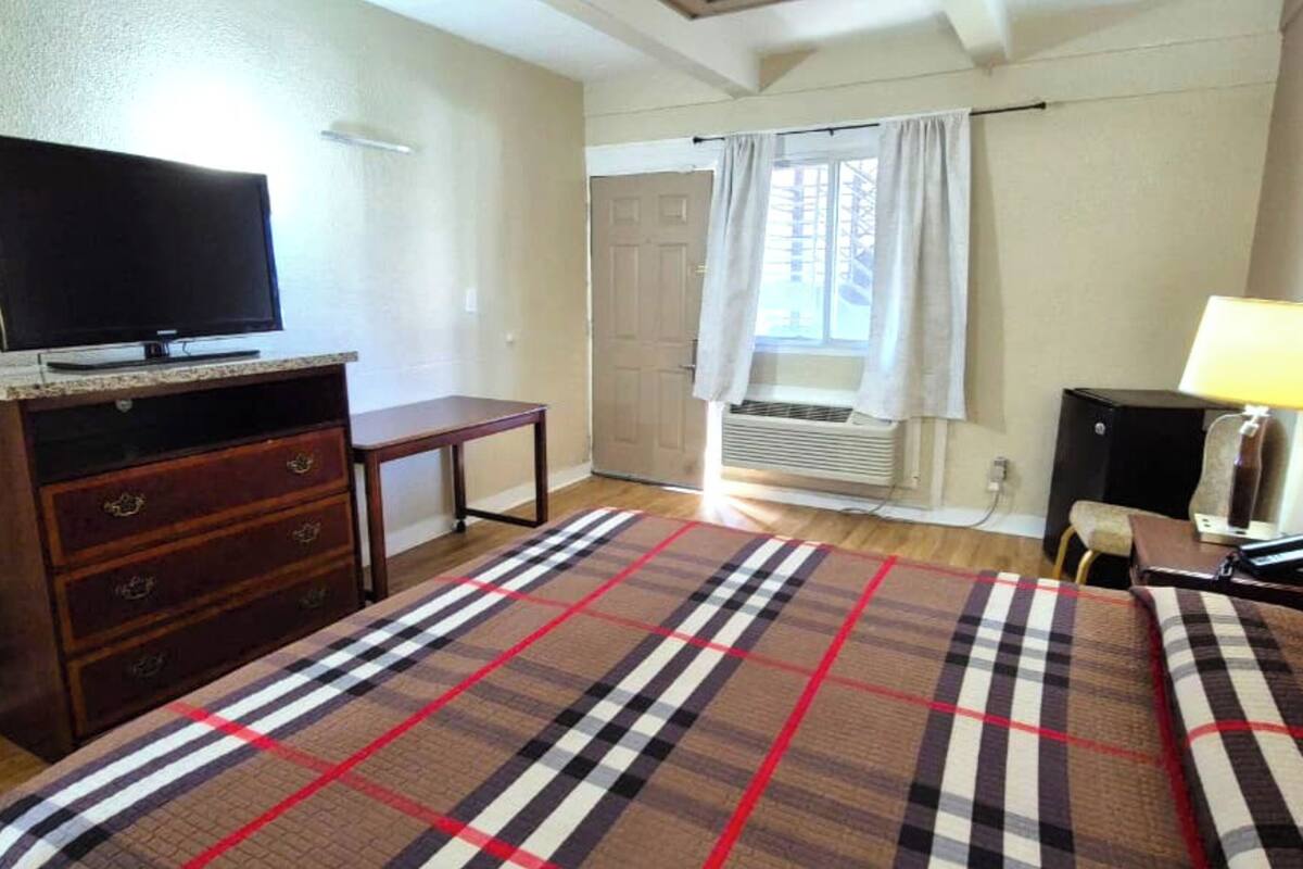 A bedroom with a bed, a television on a wooden stand, a small table, a lamp, and a window with curtains.