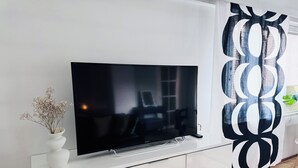Een moderne woonkamer met een flatscreen televisie op een witte kast, een vaas met gedroogde bloemen en een zwart-wit abstract gordijn.