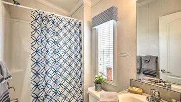 Une salle de bain avec un rideau de douche, une fenêtre avec des stores, un lavabo avec un miroir et des serviettes.