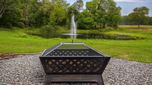 A fire pit with a metal grate, surrounded by gravel, in a grassy area with a pond and trees in the background. - The Green Forest Refuge: Cabin & Suite (Pleasant Mount)