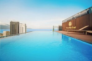 Una piscina con vistas al océano y edificios de la ciudad, una terraza de madera con sillones de descanso, y el cartel del Hotel Ocean Bliss.