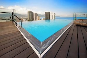 A swimming pool with a wooden deck, surrounded by high-rise buildings and a clear sky. - Ocean Bliss Hotel (Da Nang)