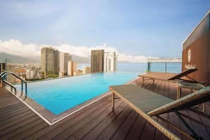 A rooftop infinity pool with wooden deck and lounge chairs, overlooking a cityscape and the ocean. - Ocean Bliss Hotel (Da Nang)