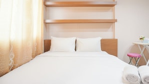 Una cama bien tendida con sábanas blancas, cabecera de madera y una mesita con un taburete rosa a un lado.