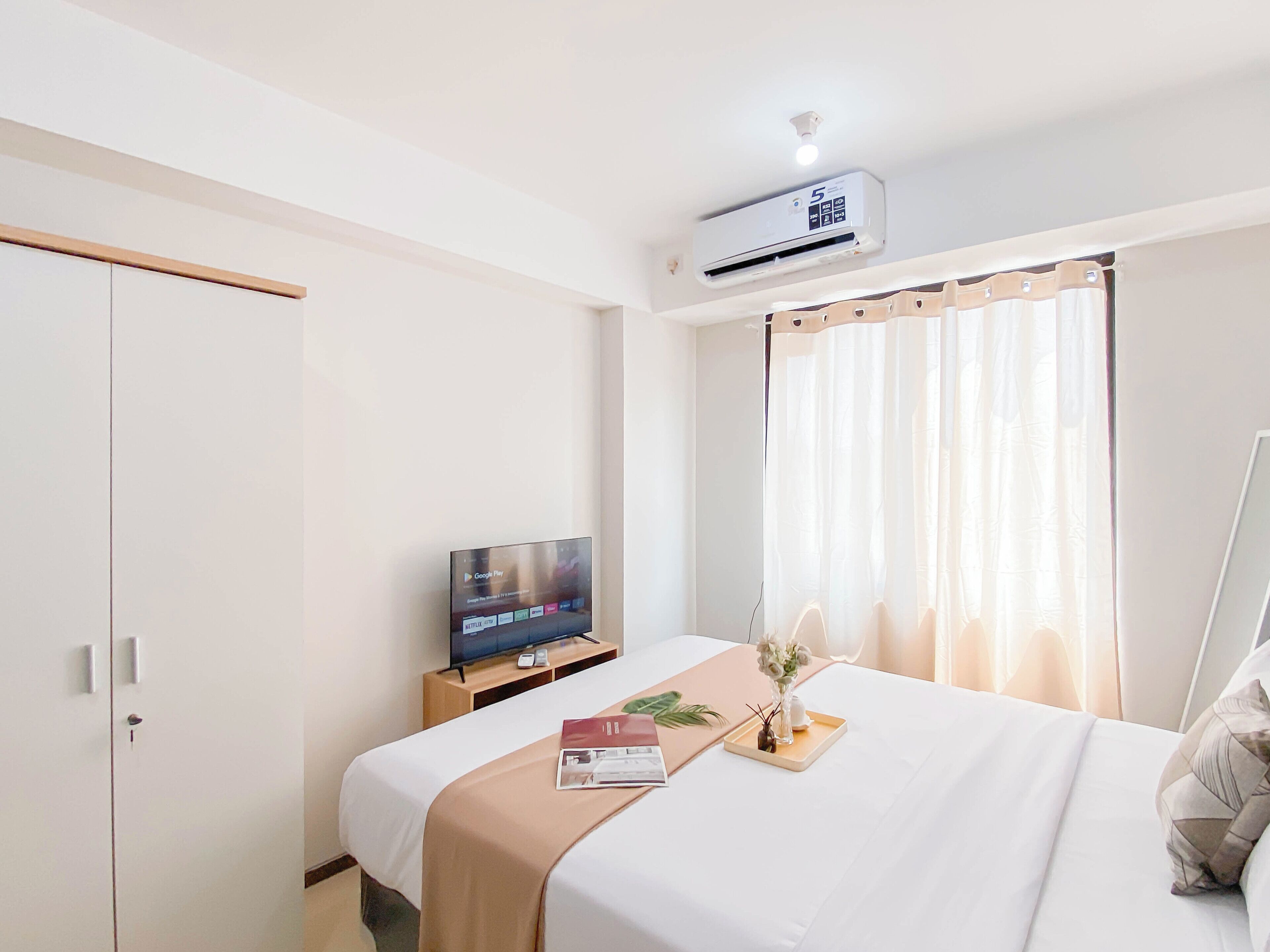 Hotellihuone, jossa on sänky, puinen TV-taso, ilmastointilaite, ikkuna, jossa on ohuet verhot, sekä yöpöytä, jossa on maljakko täynnä kukkia ja kirja.