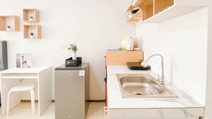 Een compact keukenhoekje met een inbouwkoelkast, een spoelbak en een klein aanrecht. Er staan planken met planten en een tafeltje met een stoel.