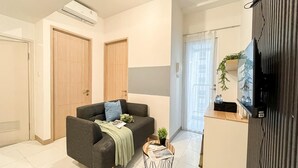 A modern living room with a sofa, a coffee table, and a TV on a stand. There are two doors and a window with curtains. - Scandinavian Touch With 2Br At Tokyo Riverside Pik 2 Apartment (Teluknaga)