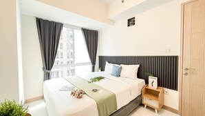 A modern bedroom with a large bed, a bedside table, a window with curtains, and a view of buildings outside. - Scandinavian Touch With 2Br At Tokyo Riverside Pik 2 Apartment (Teluknaga)