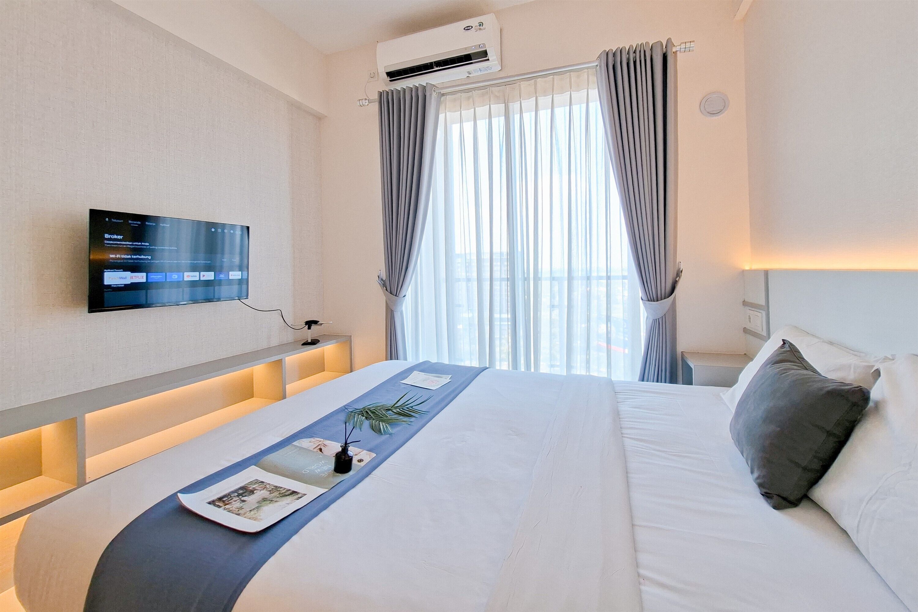 Habitación de hotel con cama, televisor de pantalla plana en la pared, mesita de noche y ventana con cortinas.