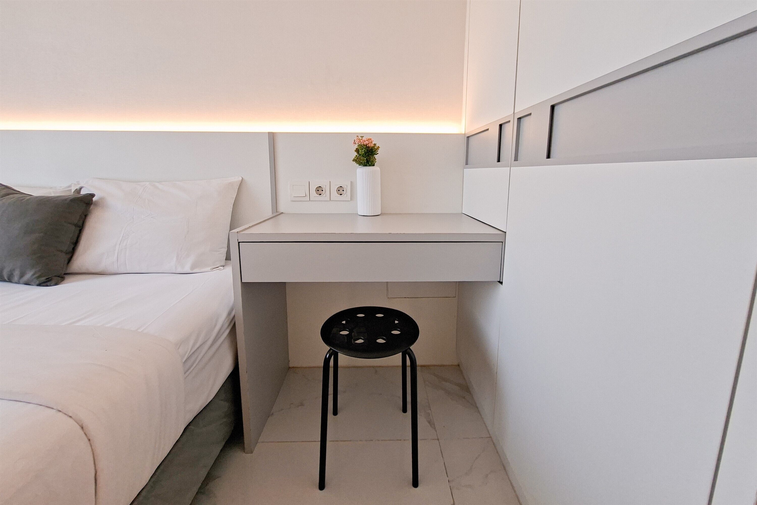 A small, modern bedroom with a bed, a desk, and a chair. There is a vase with flowers on the desk, and a wall-mounted light above the desk.