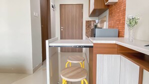 A compact kitchen with a built-in dishwasher, a small wooden stool, and a brick accent wall. - Minimalist Style Studio At Royal Sentul Park Apartment (Bogor)