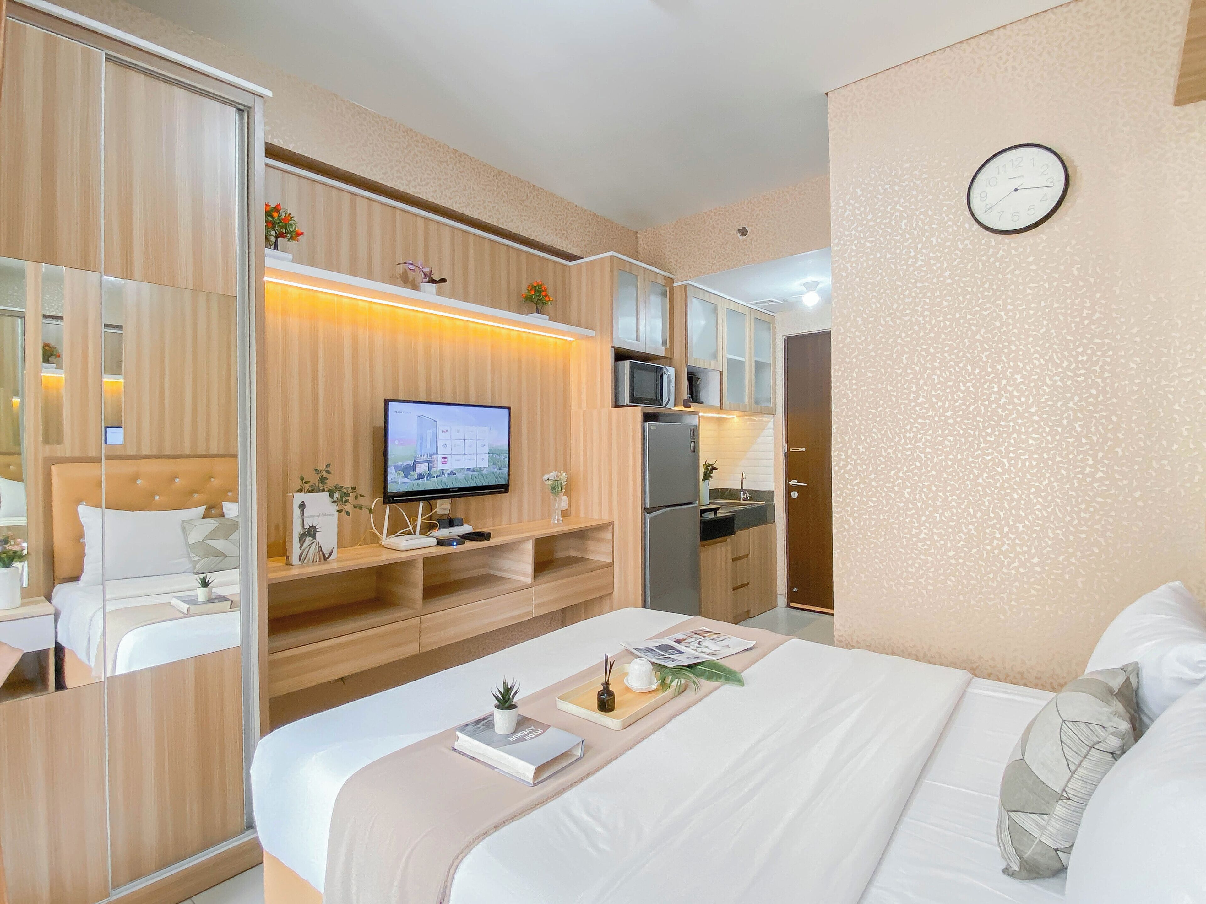 A hotel room with a bed, a TV on a wooden shelf, a refrigerator, and a clock on the wall.