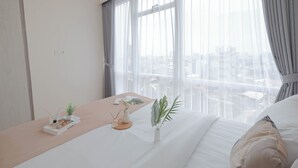 A hotel room with a bed, a bedside table with a plant, and a view of the city through sheer curtains. - Solace 2Br Near Cikini Station At Menteng Park Apartment (Jakarta)
