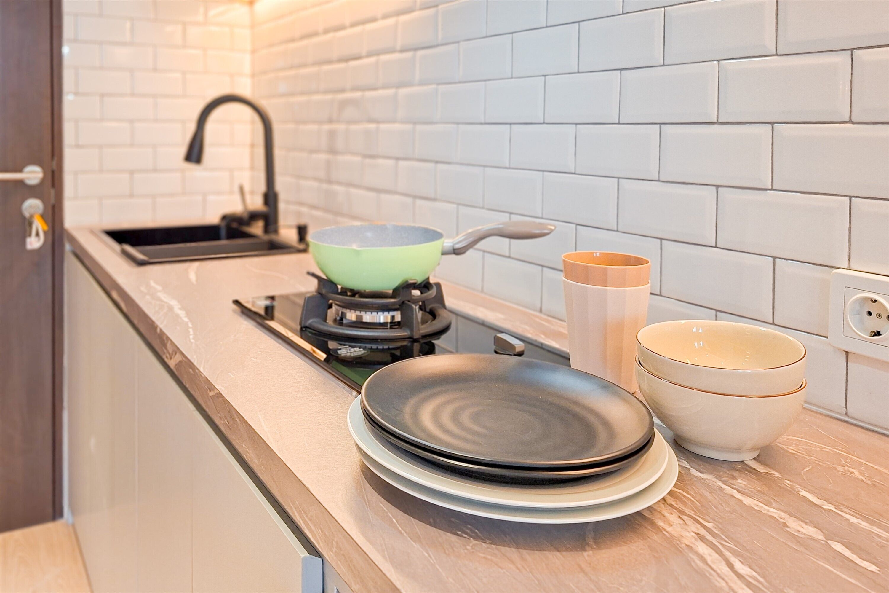 A kitchen with white tiled backsplash, a gas stove, a frying pan, a stack of plates, and a set of bowls.