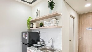 A compact kitchen with a refrigerator, sink, and countertop. There is a wooden door and a shelf with plants and kitchen items. - White Accent Studio At Apartment Meikarta Near Paramadina University (Cikarang)