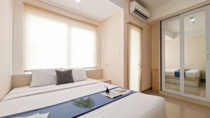 A hotel room with a bed, a bedside table, a window with blinds, an air conditioning unit, and a sliding glass door leading to another room. - Comfy And Functional Studio At Apartment B Residence (Pagedangan)