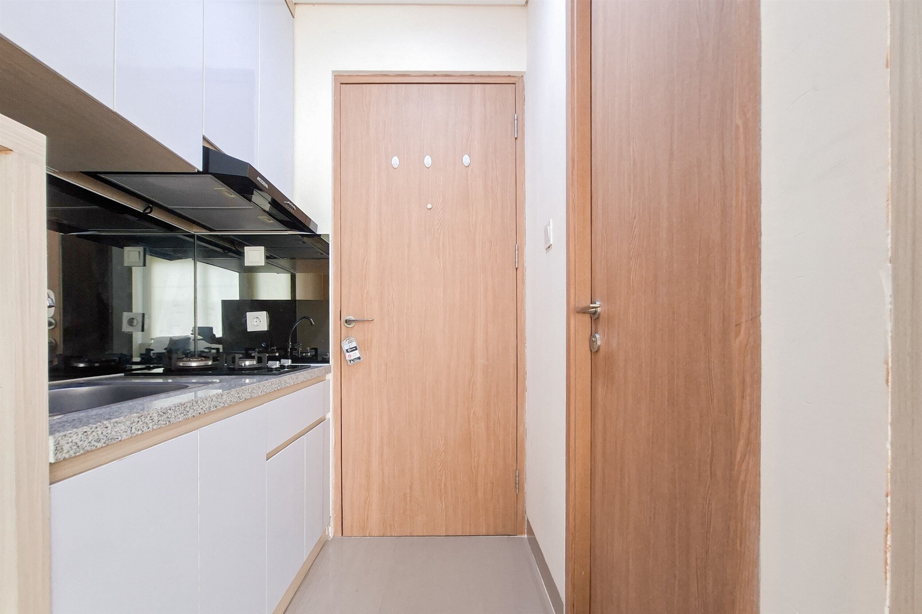 Un couloir étroit avec une porte en bois, un coin cuisine avec une cuisinière et un évier, et un meuble blanc.