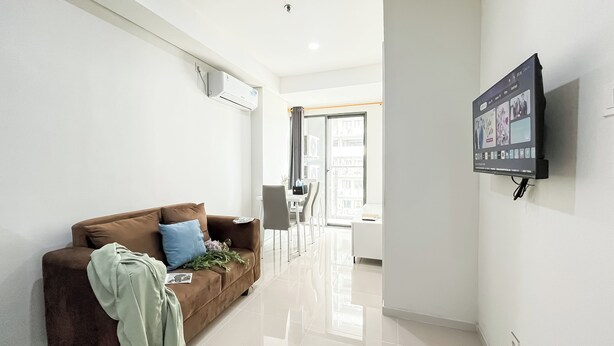 A modern living room with a brown sofa, a flat-screen TV mounted on the wall, and a dining area with a table and chairs. - Serene Stay 2Br At Daan Mogot City Apartment (Jakarta)