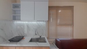 Une cuisine compacte équipée d’une cuisinière intégrée, d’un évier et d’un plan de travail. On y trouve des placards en bois sous le plan de travail et une fenêtre avec des stores sur le côté droit.