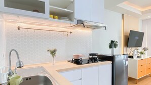A modern kitchen with a stainless steel sink, a gas stove, a built-in oven, and a refrigerator. There is a white cabinet above the sink and a white countertop. A small plant is on the countertop, and a TV is mounted on the wall. - Studio Near Rajawali Station At Amethyst Apartment (Jakarta)