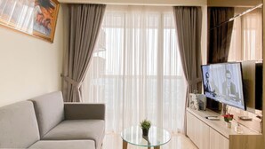 A living room with a grey sofa, a glass coffee table, a TV on a wooden cabinet, and sheer curtains. - 2Br With Private Lift Near Station At Menteng Park Apartment (Jakarta)