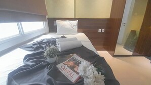 A modern hotel room with a neatly made bed, a window with blinds, a small potted plant, and a magazine on the bed. - 1Br Near Unpar At Parahyangan Residence Apartment (Bandung)