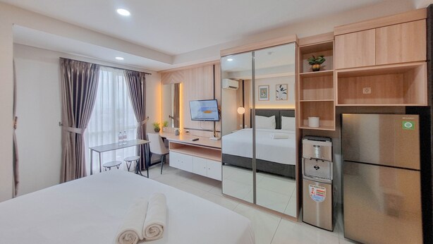 A hotel room with a bed, a desk, a TV, a mirror, a refrigerator, and a small plant. - Modern Studio Near Ambarrukmo Plaza At Barsa City Apartment (Yogyakarta)