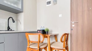 A small kitchenette with a sink and a faucet, two wooden chairs with woven seats, a small table with a potted plant and a box labeled "travelio," and a wooden door with a handle. - Soft Grey 1Br At Royal Sentul Park Apartment (Bogor)