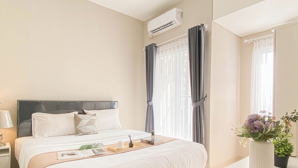 A hotel room with a bed, bedside table, lamp, air conditioning unit, curtains, and a vase with flowers. - Soft Grey 1Br At Royal Sentul Park Apartment (Bogor)
