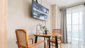 A hotel room with a television mounted on the wall displaying a streaming service interface, a round wooden table with two wicker chairs, and a window with curtains. - Studio with View at Royal Sentul Park (Bogor)