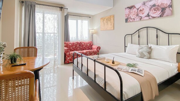 A well-lit bedroom with a bed, a red floral sofa, a wooden table with a chair, and a large window with curtains. - Studio with View at Royal Sentul Park (Bogor)