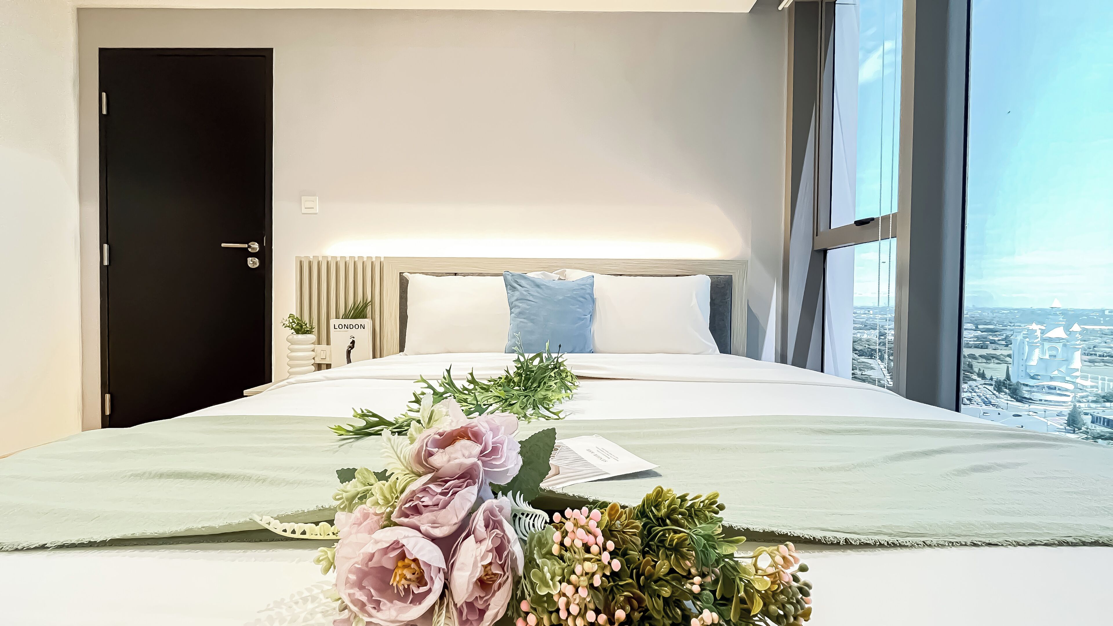 Ein Hotelzimmer mit Bett, Nachttisch, Blumentopf und Blick auf eine Stadtlandschaft durch große Fenster.