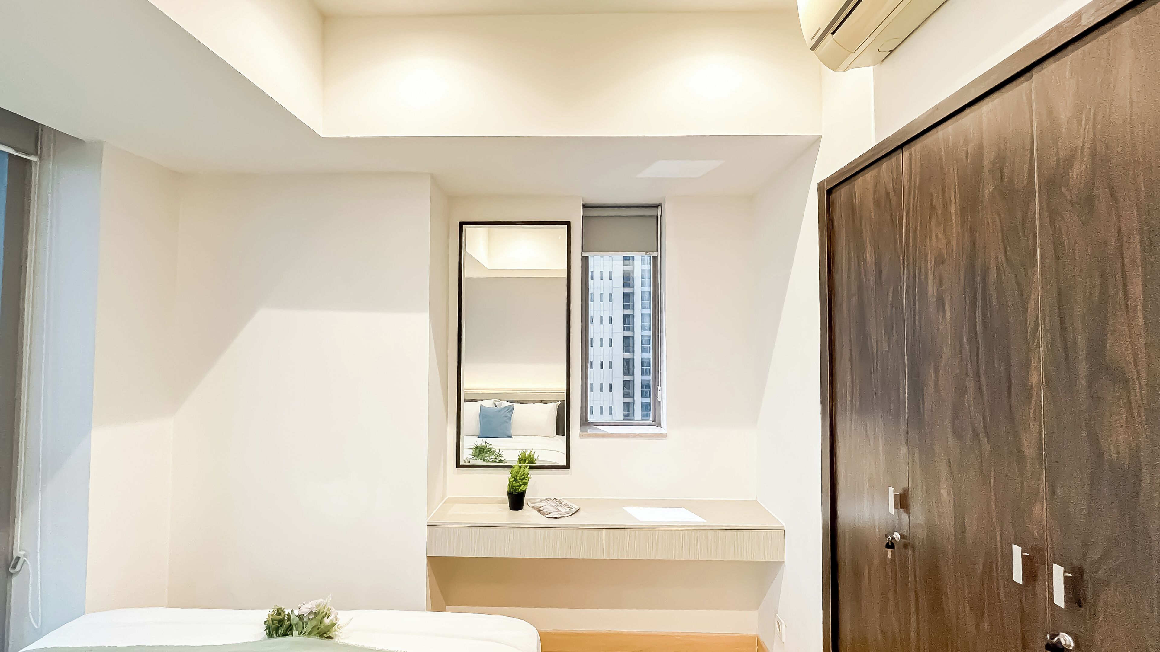 A modern bedroom with a large mirror, a small window, a wooden wardrobe, and a bedside plant.