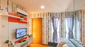 A living room with a sofa, a TV on a stand, a wall-mounted air conditioner, and a door with curtains. - 2Br Near Mall At Cinere Bellevue Suites Apartment (Depok)
