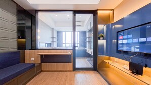 Una habitación de hotel moderna con suelo de madera, zona de estar con un banco, un televisor de pantalla plana grande montado en la pared y una puerta de cristal que da a un balcón.