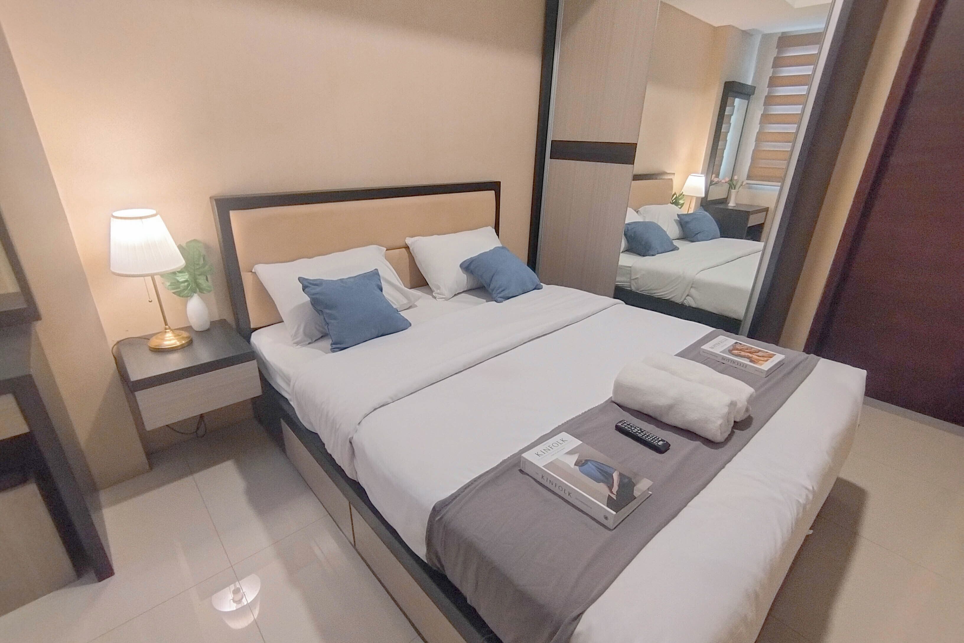 A neatly made bed with pillows, a bedside lamp, a remote control, and a book on the bed.