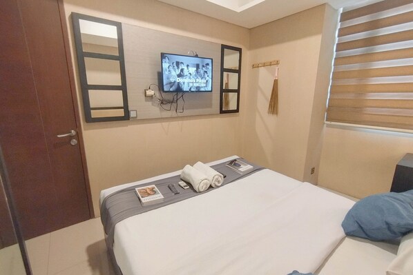 Habitación de hotel con cama, televisor montado en la pared, ventana con persianas y puerta.