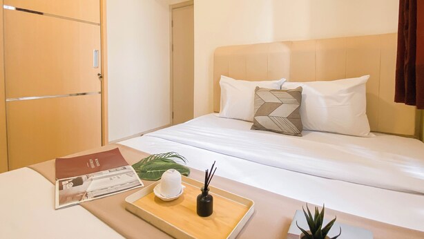 A hotel room with a bed, a bedside table with a tray, a plant, and an air conditioning unit. - Basic 2Br Apartment At Meikarta Near Dian Harapan School (Cikarang)
