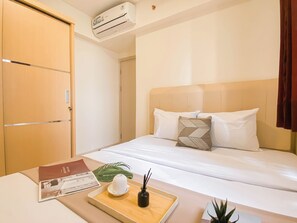 A hotel room with a bed, a bedside table with a tray, a plant, and an air conditioning unit. - Basic 2Br Apartment At Meikarta Near Dian Harapan School (Cikarang)