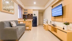 A modern living room with a grey sofa, a small dining table, and a kitchen area with a fridge, microwave, and sink. - Basic 2Br Apartment At Meikarta Near Dian Harapan School (Cikarang)