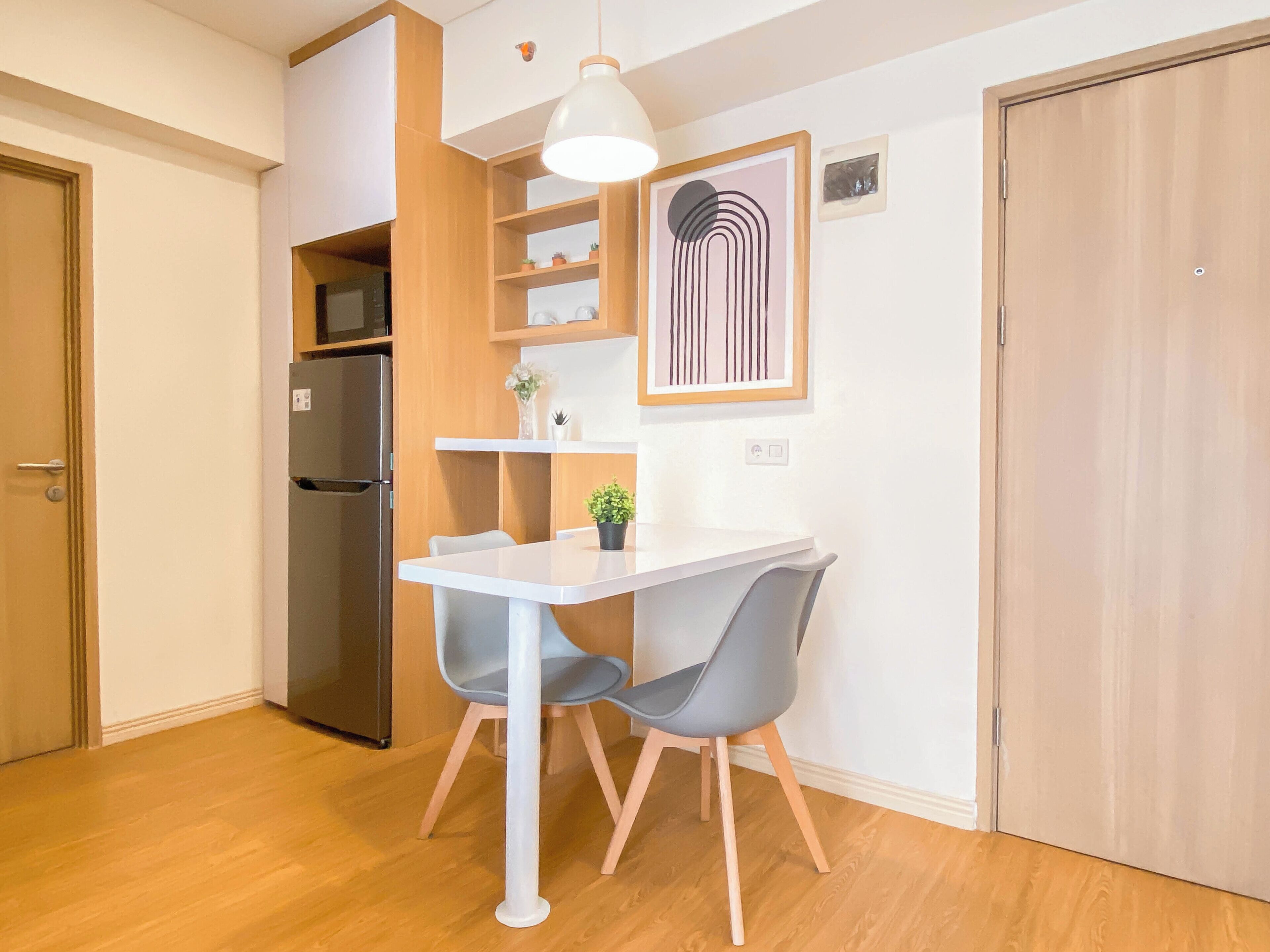 A compact kitchen with a white table, two grey chairs, a hanging pendant light, a refrigerator, and a wooden door.